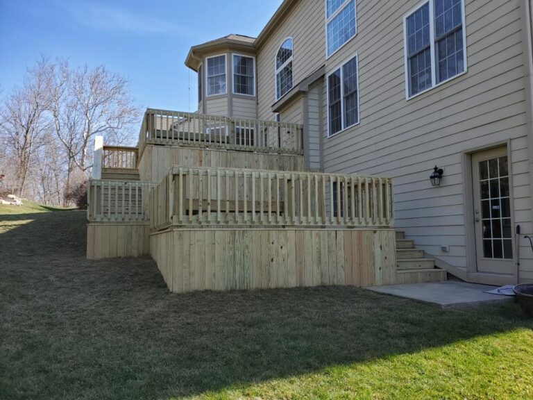 "Multi-level wooden deck extending from a beige house, surrounded by grass and trees."