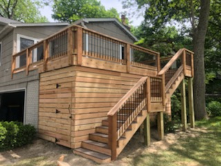 "Wooden deck and staircase with railing, attached to a house and surrounded by greenery."