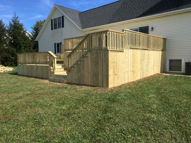 Wooden deck with railings and stairs attached to a house, surrounded by grass and trees under a blue sky.
