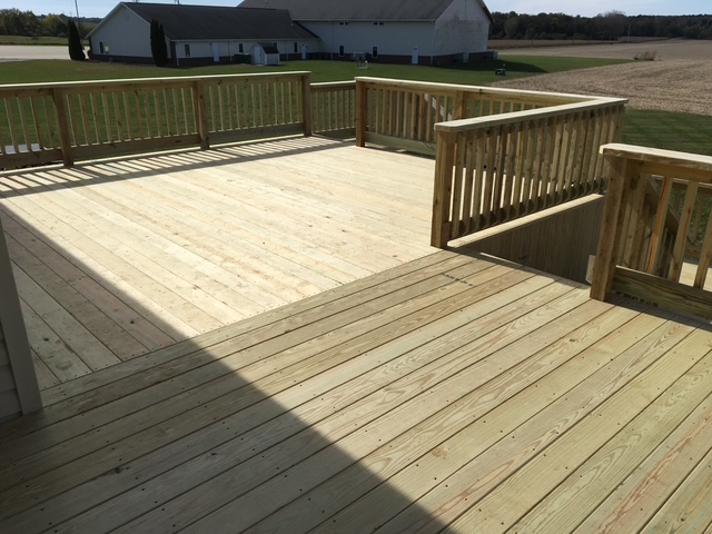 A large wooden deck with railings, showcasing freshly constructed planks and an expansive grassy field in the background.