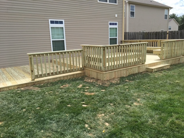 Wooden deck in a backyard, featuring railings and a multi-level design, adjacent to a gray house with white windows.