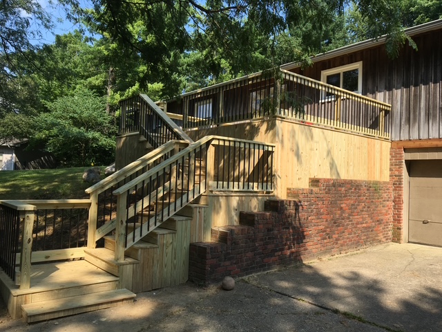 "Wooden deck with black railings and a connecting staircase at a residential home surrounded by greenery."