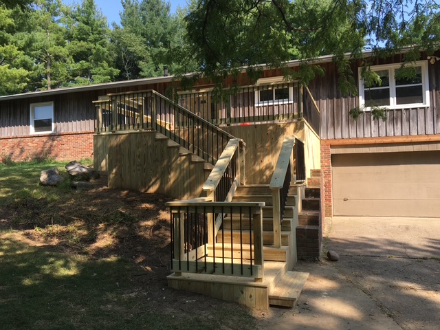 A wooden walkway with stairs leading to a house, surrounded by greenery, featuring a garage in the background.