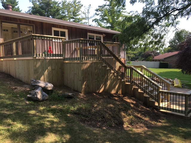 "Deck with stairs leading to a wooden house, surrounded by greenery and rocks."