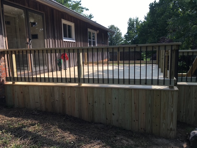 "View of a wooden deck with black railings in front of a house surrounded by trees."