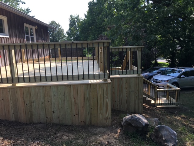 A newly constructed wooden deck with railing, adjacent to a house, featuring a staircase and surrounding greenery.