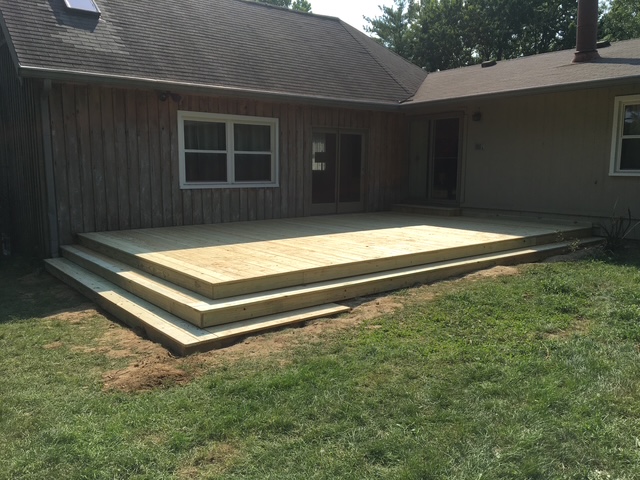 Newly constructed wooden deck with steps, extending from a house back entrance, set on a grassy area.