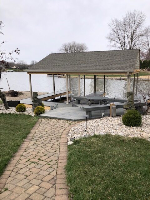 Dock with a covered seating area by a lake, surrounded by grass and stone landscaping, leading to a paved walkway.