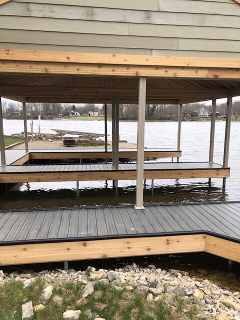Image of a lakeside pavilion structure with wooden decking and posts above water, surrounded by grass and a scenic view of the lake.