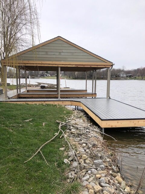 "Wooden dock with a covered seating area extending over a lake, surrounded by grass and rocky shoreline."