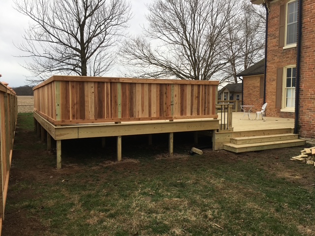 "Wooden deck with railing attached to a brick house, featuring a staircase and furniture on a grassy yard."