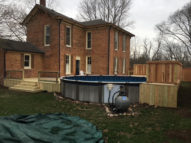 A two-story brick house with a circular above-ground pool, wooden deck, and landscaping in the backyard.