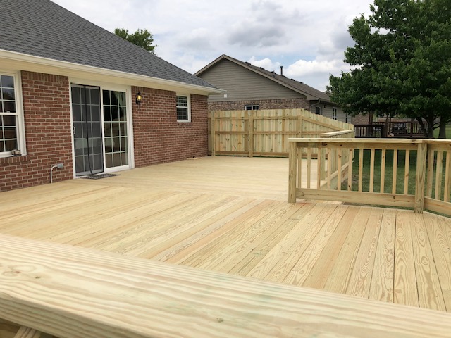 Newly renovated wooden deck extending from a brick house, surrounded by fencing and green lawn under a cloudy sky.