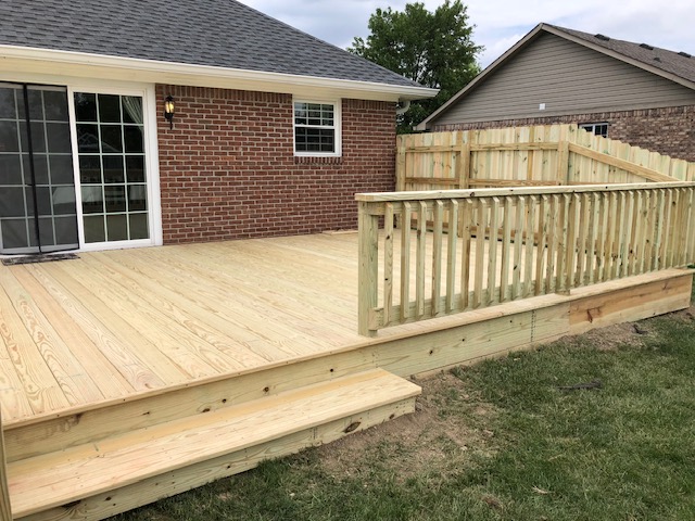 Newly constructed wooden deck with railings and steps, adjacent to a brick house and grass lawn.