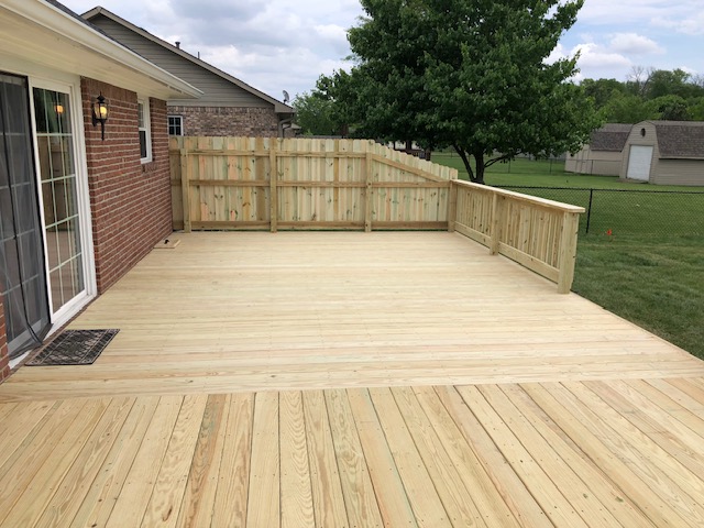 Newly constructed wooden deck with a fenced perimeter adjacent to a brick house, surrounded by green grass and trees.