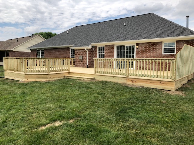 Newly constructed wooden deck with railing extending from the back of a brick house, surrounded by grass and under a partly cloudy sky.