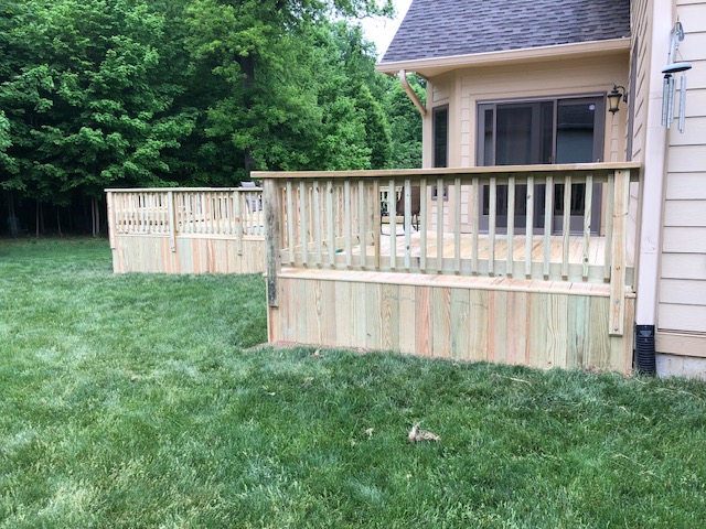 "Wooden deck with railings adjacent to a house, surrounded by lush green grass and trees."