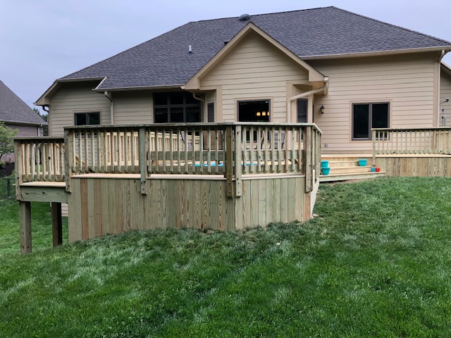 A modern house with a spacious wooden deck and grassy yard, showcasing a neutral-colored exterior under a cloudy sky.