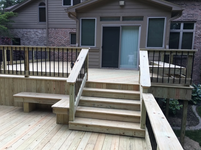 Wooden deck with stairs leading to a glass door entry, featuring black railings and a spacious design in a residential backyard setting.