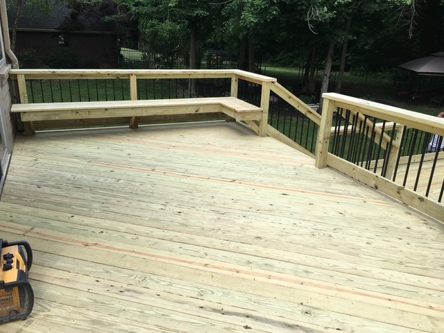 A newly constructed wooden deck featuring a railing and built-in bench seating, surrounded by greenery.