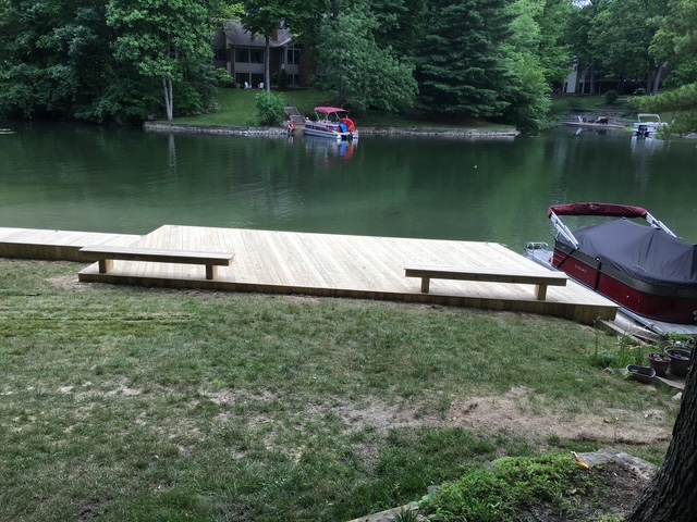 Wooden dock overlooking a serene lake with boats moored in the background and lush greenery surrounding the area.