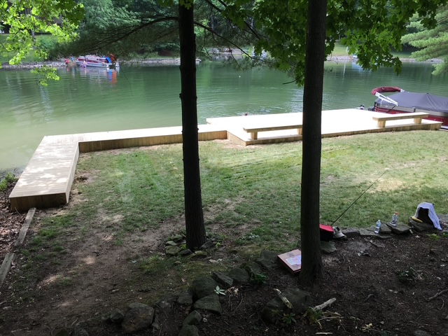 "Wooden deck by a tranquil lake, surrounded by grass and trees, with boats visible in the background."