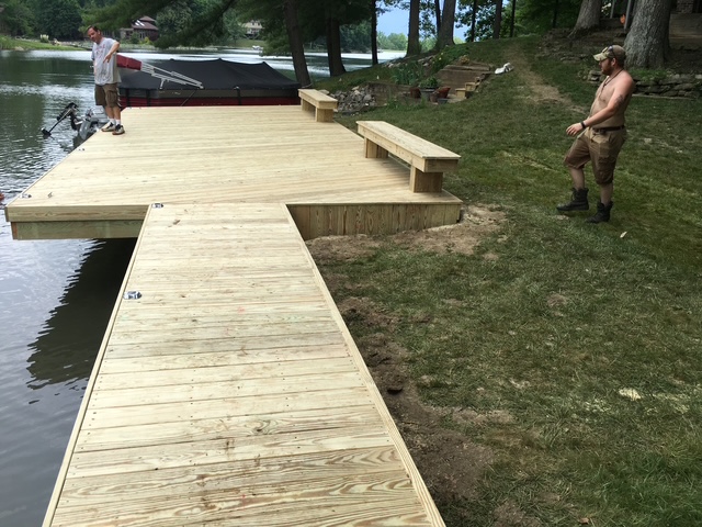 "Two men work on a wooden dock by the water, with one standing on the dock and the other on the grass nearby, amidst a forested backdrop."