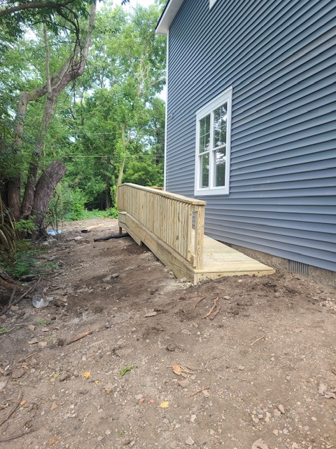 "Wooden ramp leading to a side entrance of a gray house, surrounded by a gravel and soil landscape."