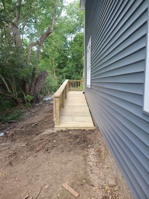 A newly constructed wooden ramp alongside a gray house, leading to a wooded area, with unpaved ground nearby.