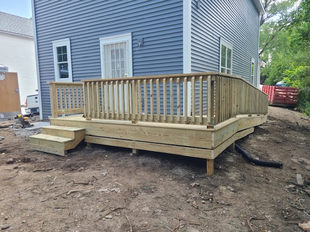 A newly built wooden deck with railings and steps, situated next to a blue house and surrounded by a clearing.