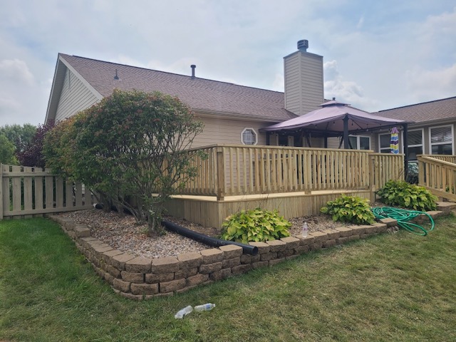 A wooden deck with a pergola, surrounded by landscaped stones and greenery, attached to a house under a cloudy sky.