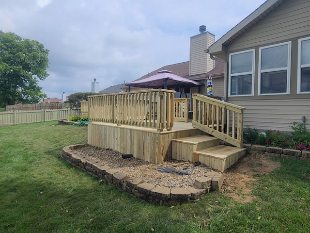 "Wooden deck with railing and steps in a backyard, surrounded by landscaped areas and a patio umbrella."