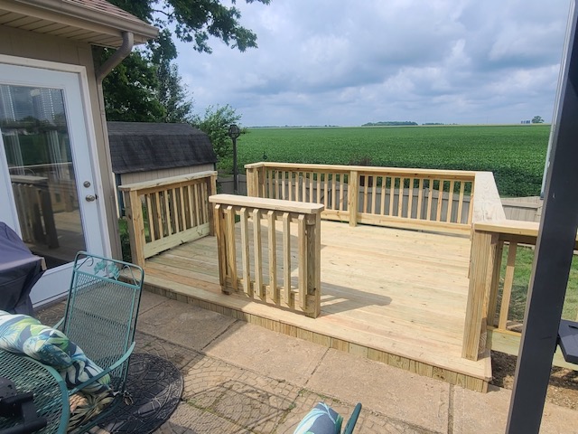 "Newly constructed wooden deck with railings overlooking a green field, adjacent to a home patio with outdoor seating."