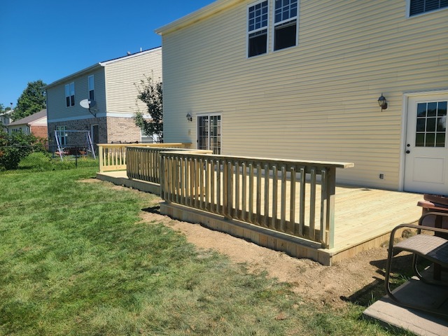 Newly built wooden deck with railing, adjacent to a light-colored house, set in a grassy yard on a sunny day.