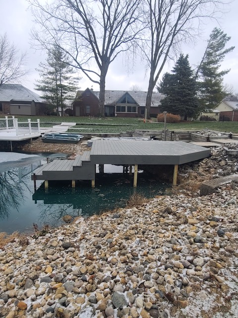 A wooden dock extends over a calm water body, surrounded by rocky shoreline and residential properties, under overcast skies.