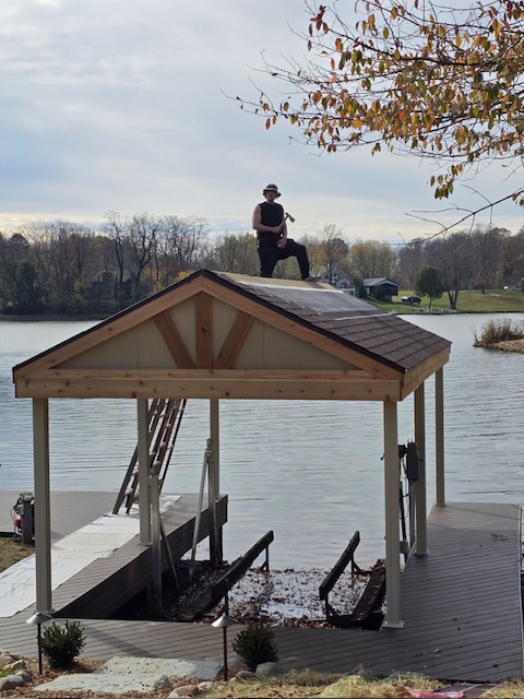 A person in black clothing sits on the roof of a lakeside gazebo, with a calm body of water and trees in the background.