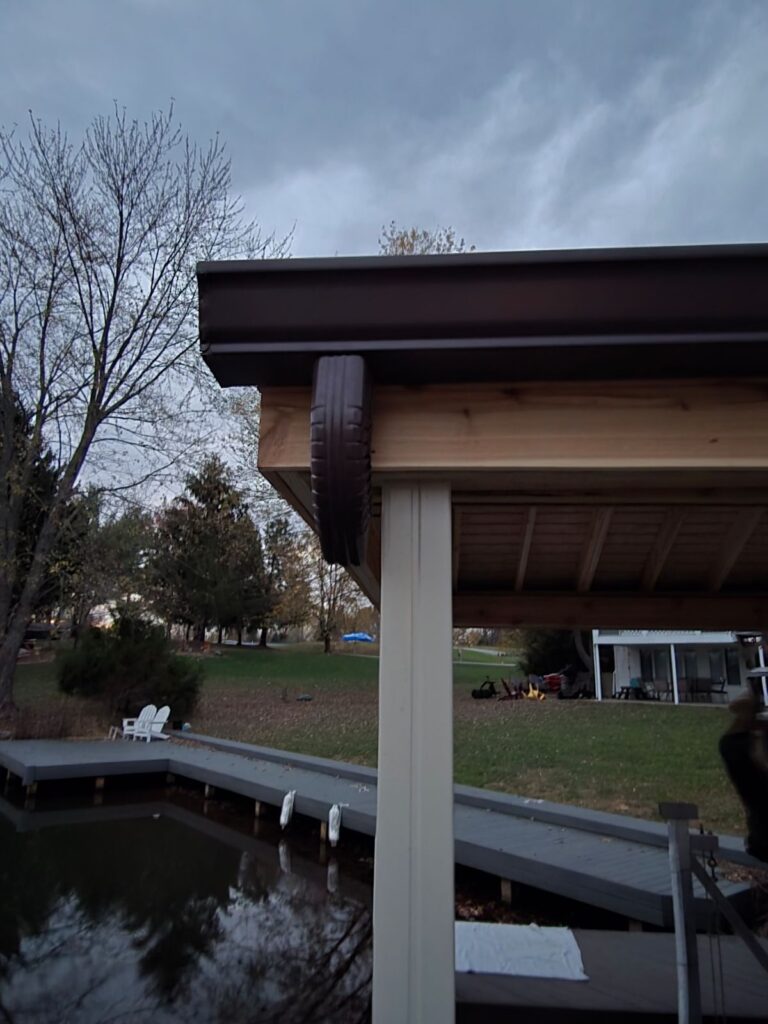 Close-up of a wooden gazebo with a dark metal roof and a unique tire feature, overlooking a tranquil pond and grassy area.