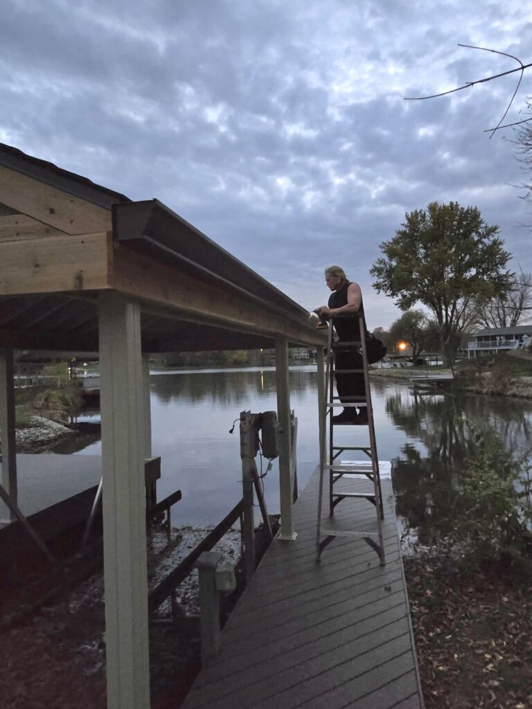Person standing on a ladder working on the roof of a dock at dusk, with calm water and cloudy skies in the background.