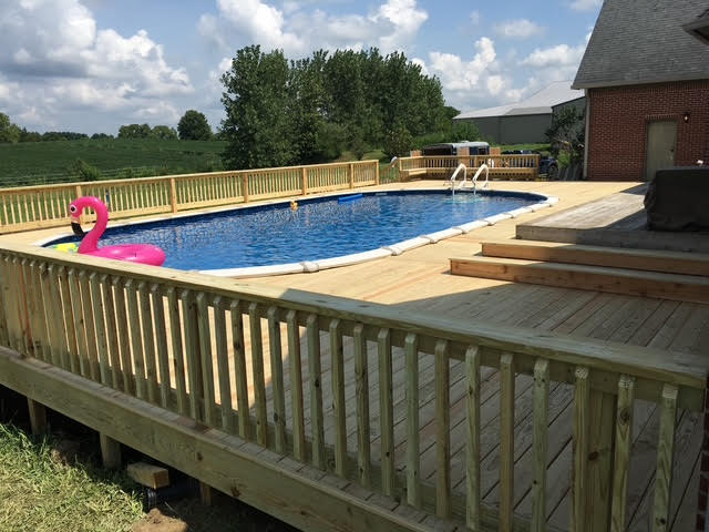 A well-maintained wooden deck surrounds a blue swimming pool, complete with a pink inflatable float and lush green trees in the background.