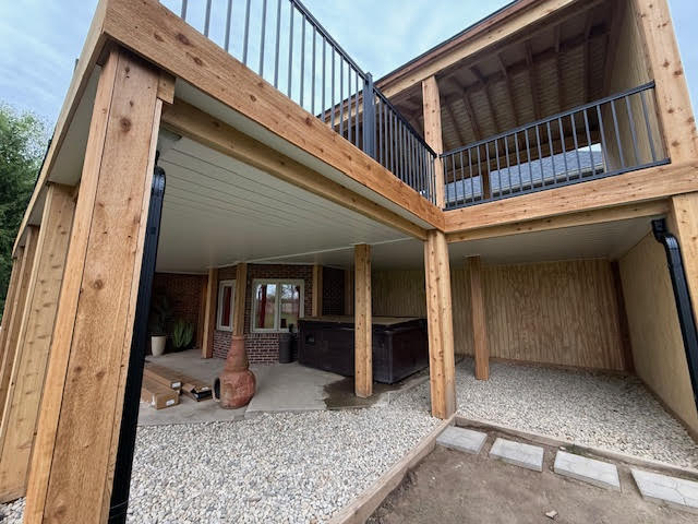 A partially constructed outdoor space featuring wooden support beams, a gravel floor, a hot tub, and a balcony above.